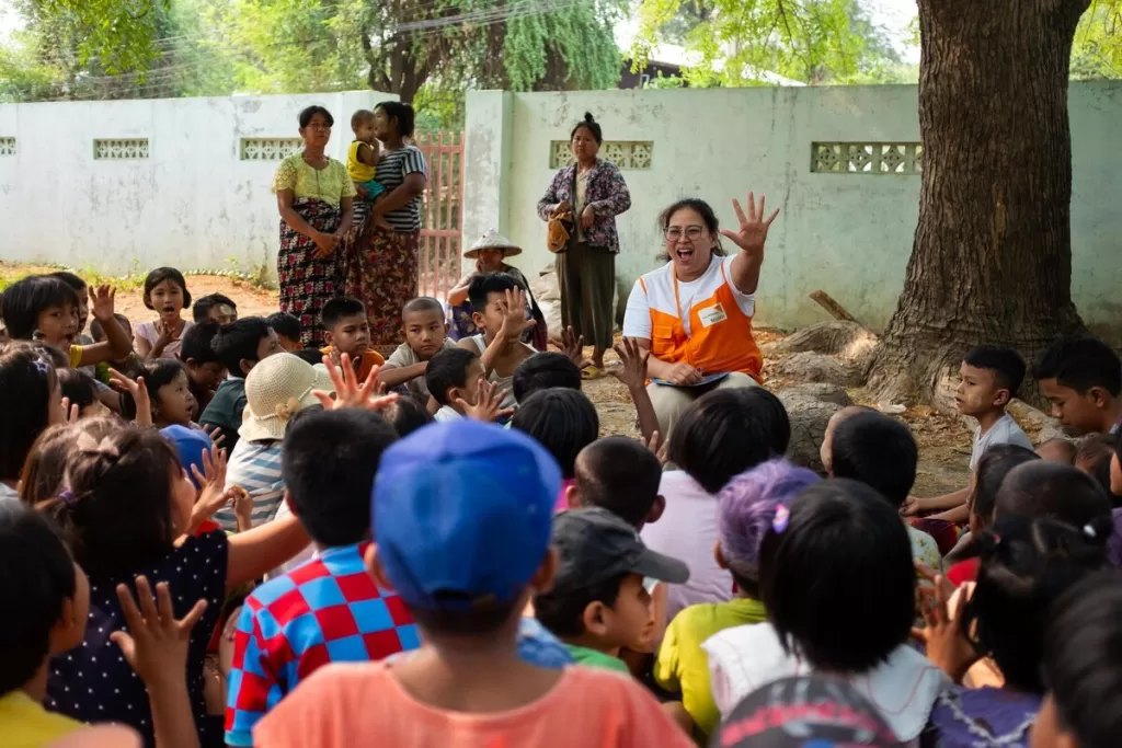 Humanitarian worker engages a large group of children in an outdoor activity, raising her hand as the children eagerly respond